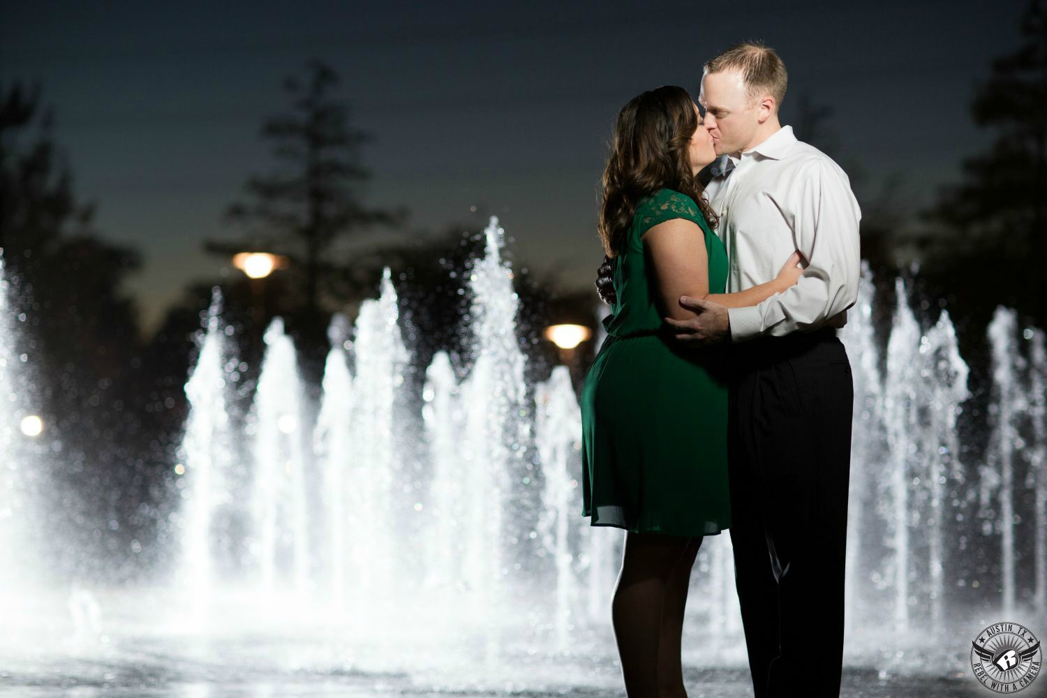 Curly haired brunette girl with a green dress with lace short sleeves embraces a sandy haired guy wearing a white button up collared shirt and black pants while kissing in front of backlit jets of water coming from the ground at the Liz Carpenter Fountain at Butler Park at dusk with dark blue twilight sky in the background in this strobe influenced engagement session in Austin.